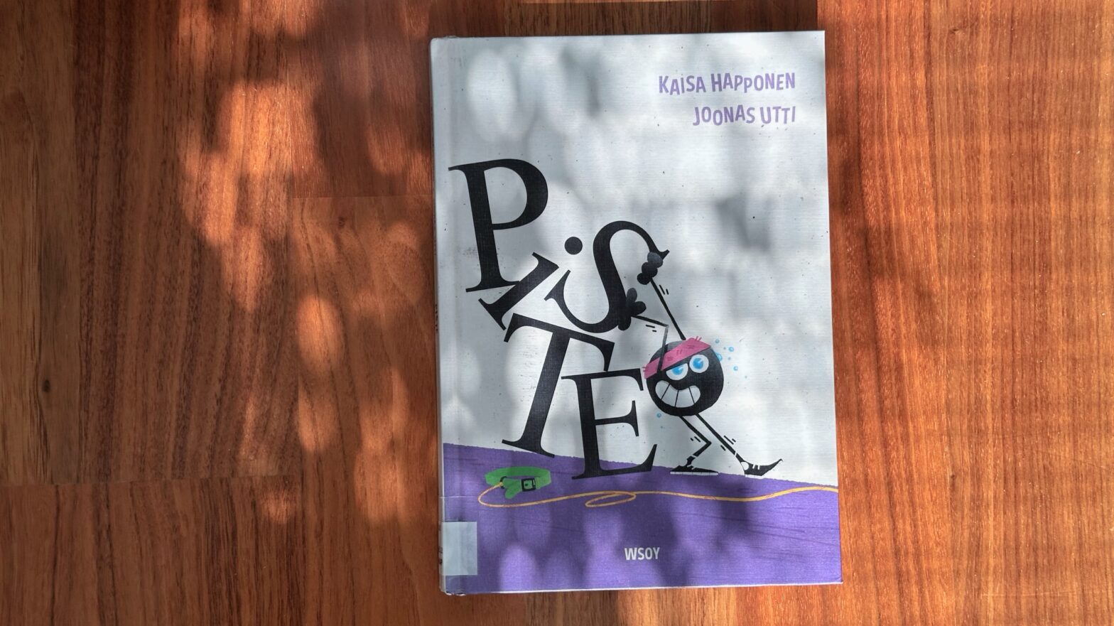 Piste-kirja lattialla sälekaihdinten ja viherkasvin auringonvalosta muodostamassa varjokuviossa.