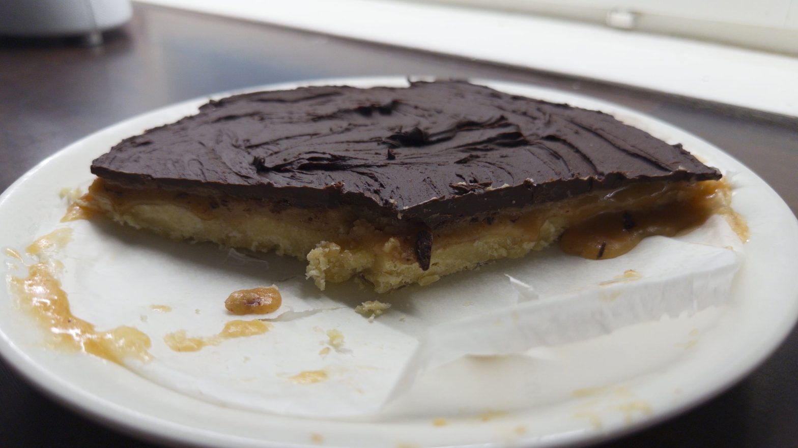 Millionaire's Shortbread eli matala leivonnainen, jossa on murotaikinapohja, kinuskikerros ja tumma suklaakuorrute päällä. Pyöreästä leipomuksesta on syöty jo noin 2/3.
